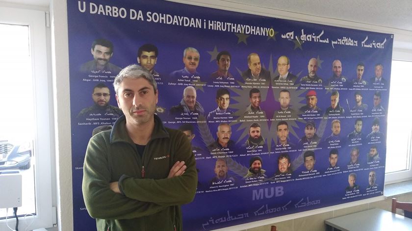 Johan Cosar em frente a um cartaz com as caras e os nomes de membros do Conselho Militar Siríaco mortos em combate. Foto: Filipe d